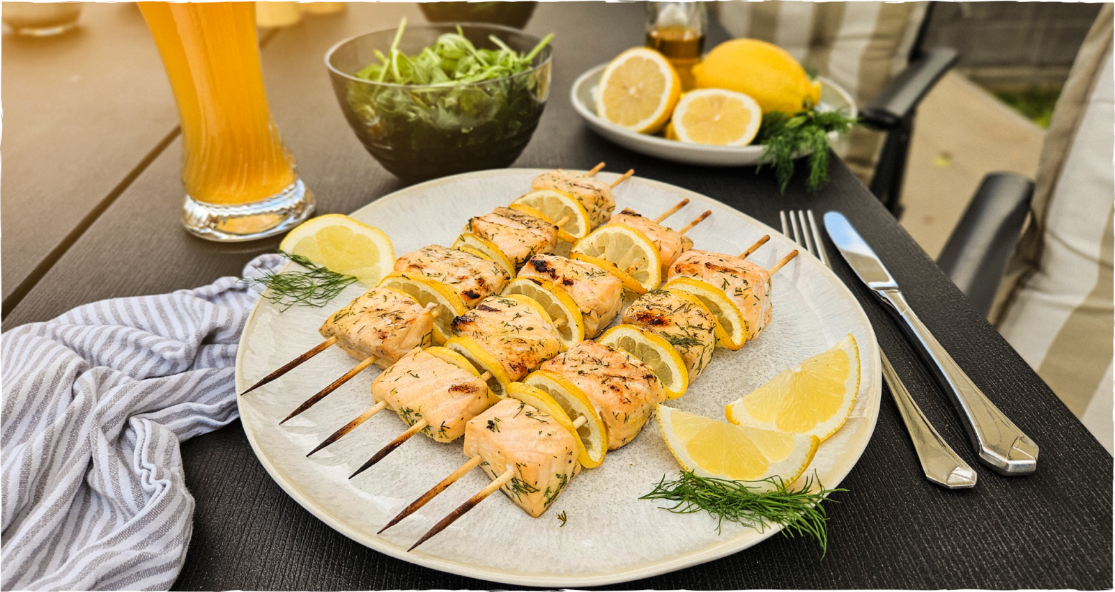 Gegrillte Lachsspieße mit Zitronenscheiben auf einem weißen Teller, serviert mit frischem Dill, Zitronenspalten, Rucolasalat und einem Glas Kapuziner Alkoholfrei.