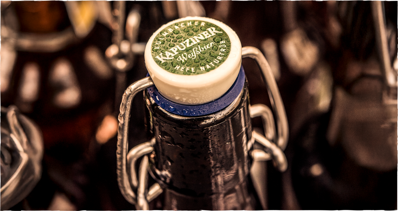 Nahaufnahme einer verschlossenen Bierflasche mit Bügelverschluss, deren Deckel das Logo der Marke Kapuziner Weißbier zeigt, mit Wassertropfen auf der Oberfläche
