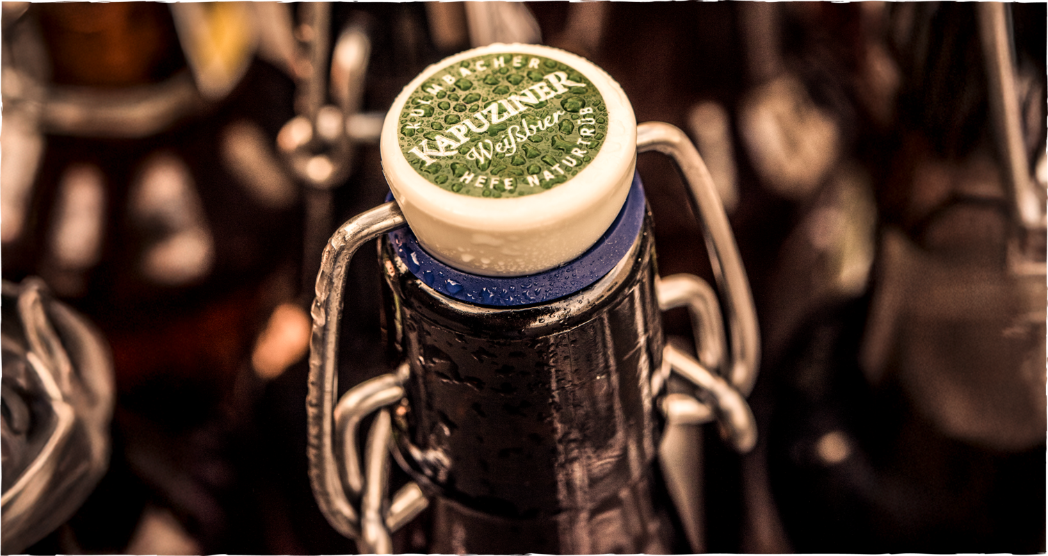 Nahaufnahme einer verschlossenen Bierflasche mit Bügelverschluss, deren Deckel das Logo der Marke Kapuziner Weißbier zeigt, mit Wassertropfen auf der Oberfläche