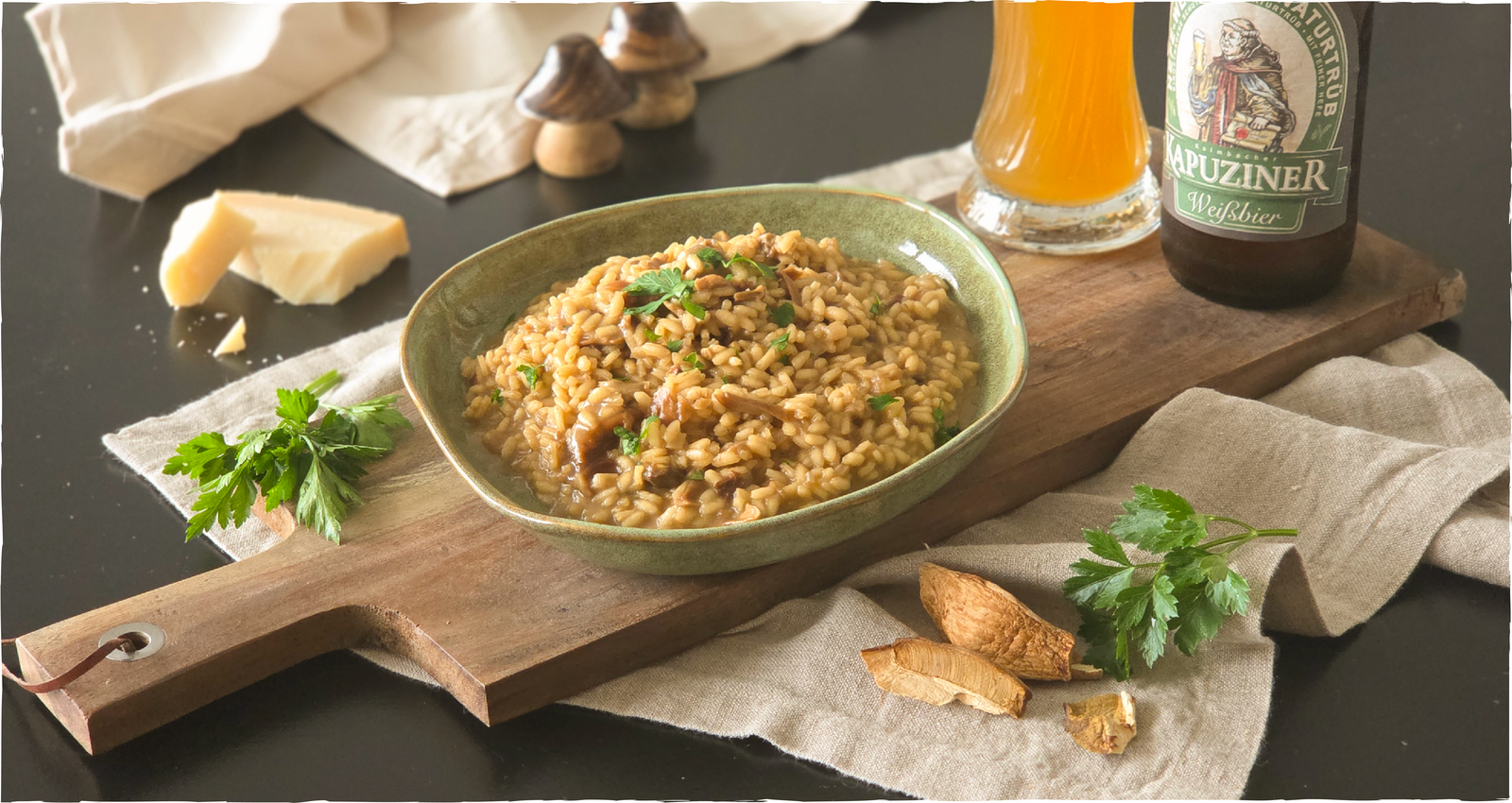 Herbstliches Steinpilzrisotto mit Petersilie in grüner Schale, dazu Parmesan; im Hintergrund Kapuziner Weißbier in Flasche und Glas