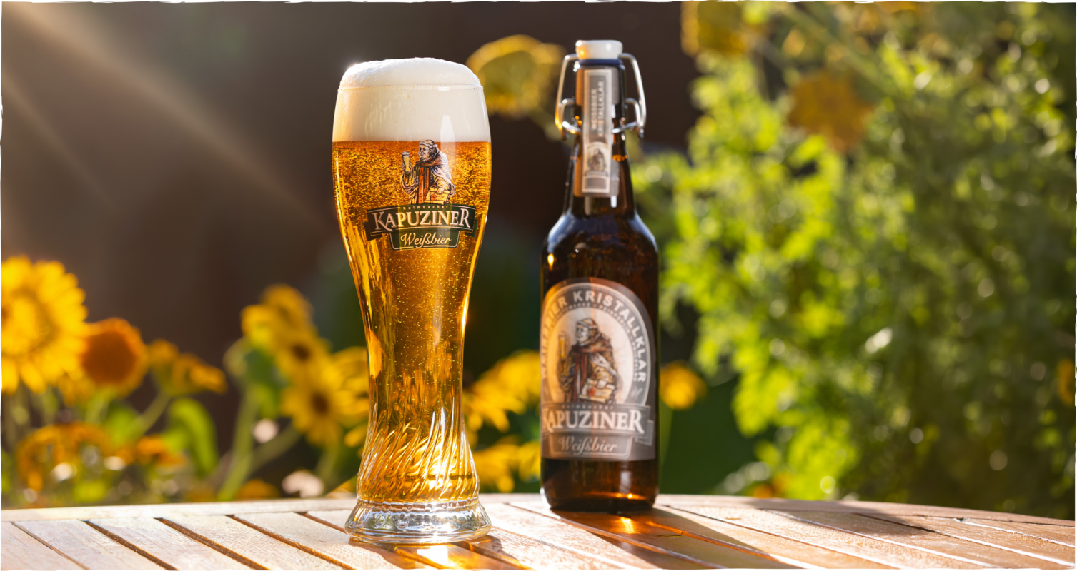 Kapuziner Weißbier Kristallweizen im Weizenglas mit dichter Schaumkrone neben Bügelverschlussflasche auf einem Holztisch im sonnigen Garten