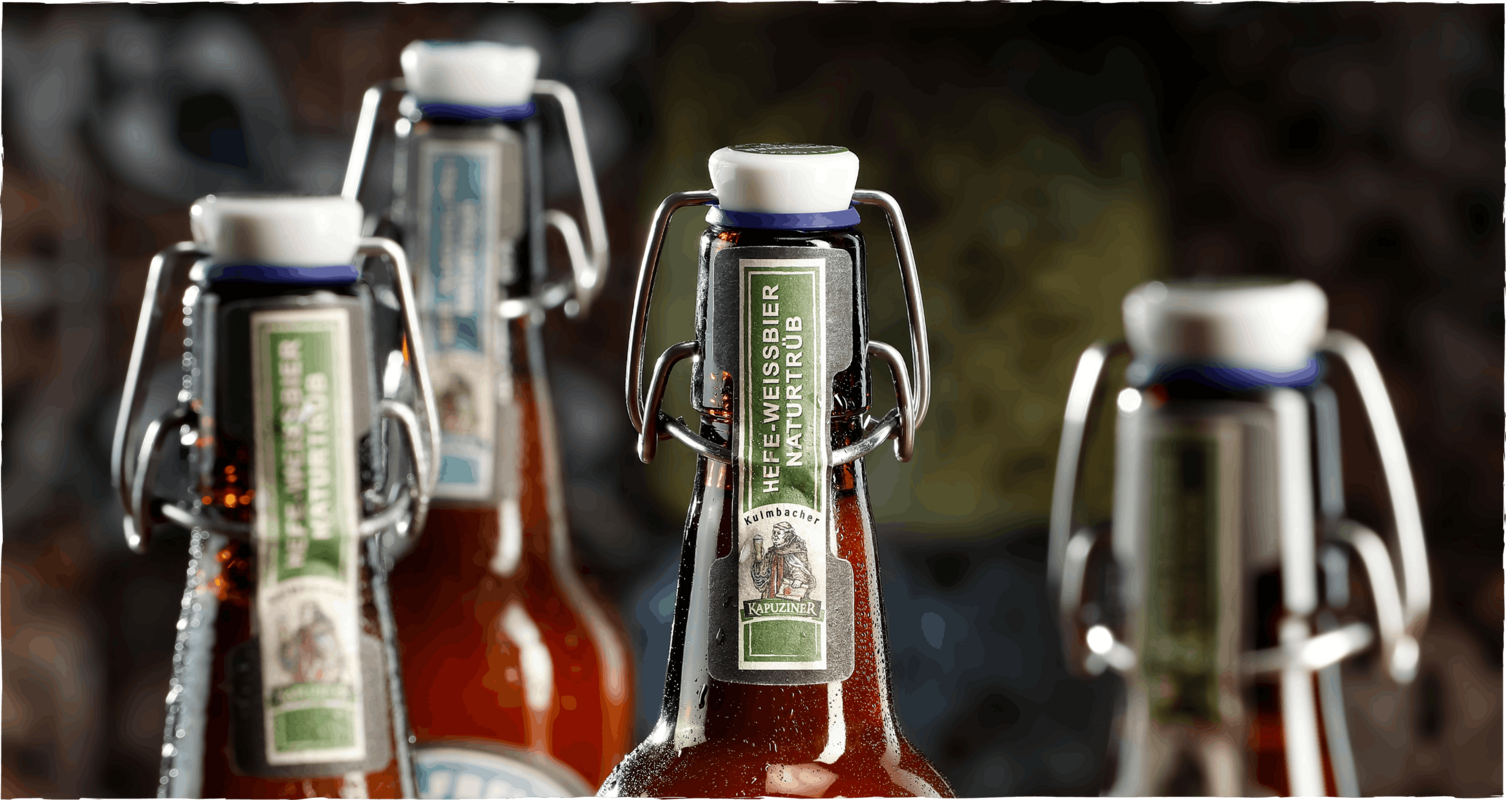 Kapuziner Weißbier Naturtrüb in Bügelflaschen mit Wassertropfen, warm ausgeleuchtet vor unscharfem Hintergrund in Brauerei-Atmosphäre.