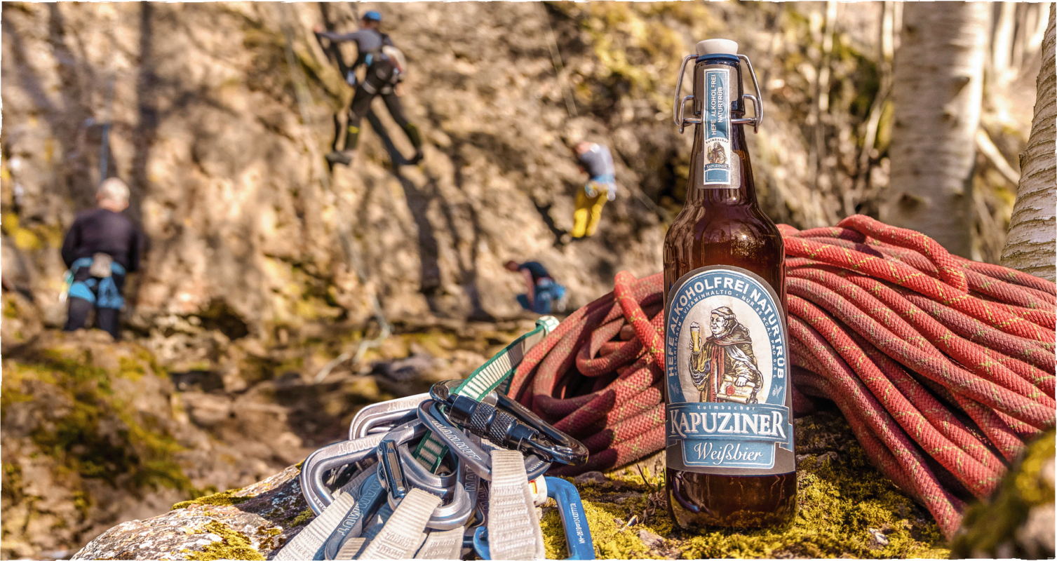 Alkoholfreies Weißbier von Kapuziner steht auf einem Felsen neben Kletterausrüstung, während im Hintergrund mehrere Personen an einer Felswand klettern